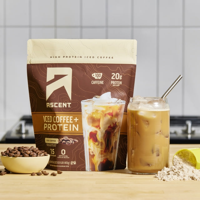 iced coffee plus protein powder in a fifteen serving size bag sitting next to a glass of iced coffee and coffee beans