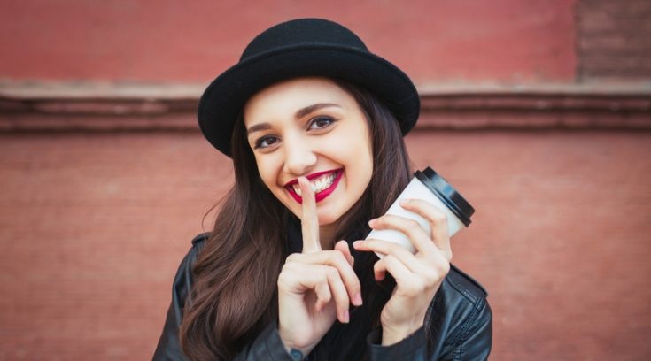 Image of smiling young woman in black bowler hat holding a coffee cup and motioning to be quiet.