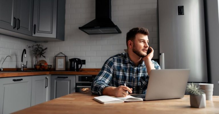 Working at Home Pros and Cons - picture of man sitting at kitchen table with laptop talking on cell phone